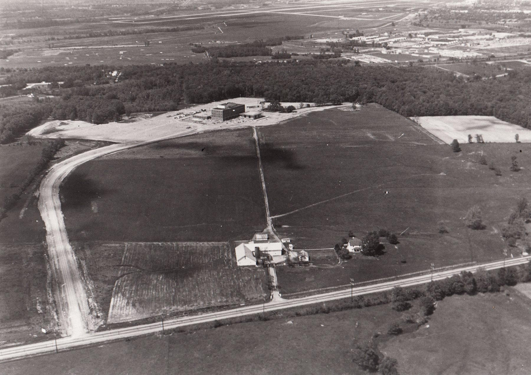 Aerial view of Dayton Campus (now WSU), showing Airway Road and the Warner farm (foreground), the "Dayton Campus" sign (lower left), Allyn Hall (top center), and a portion of Wright-Patterson AFB (upper right). (University Archives)