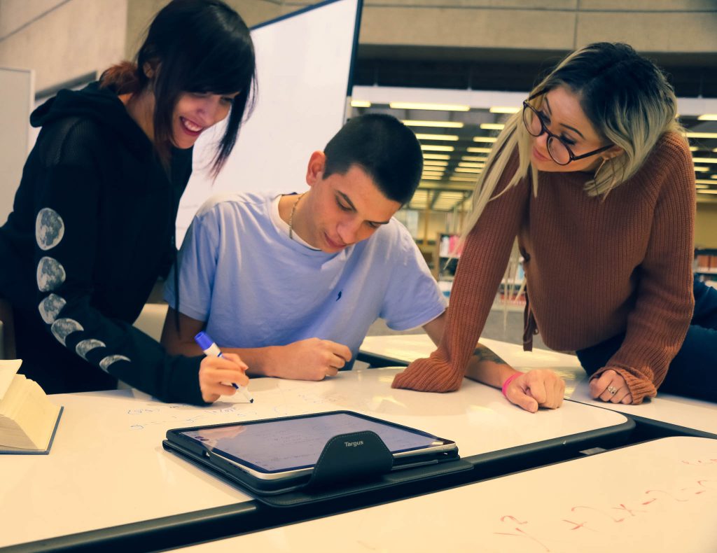 Three library users collaborate on dry erase tables with a tablet computer.